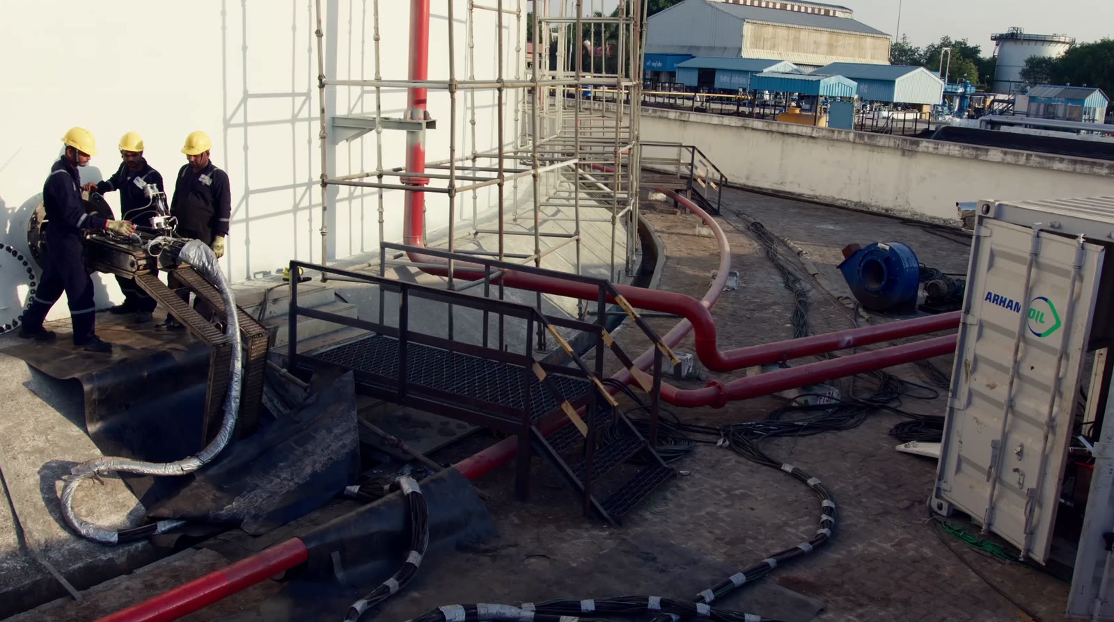 Crew in hard hats connecting hoses beside a storage tank, with an Arham Oil robotic tank cleaning container on site.