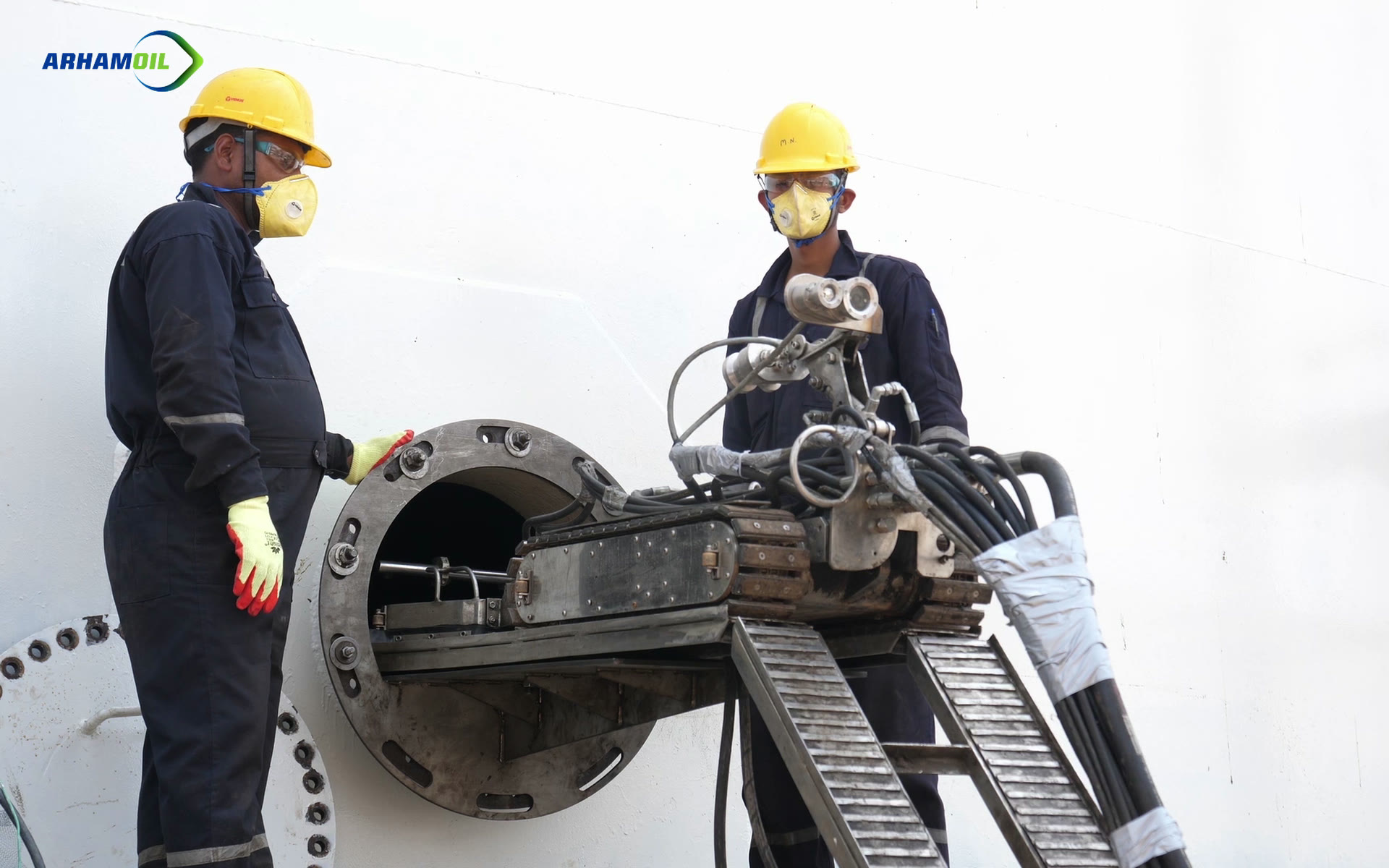 Two crew members in protective gear guide a tracked robot into a storage tank through its manway.